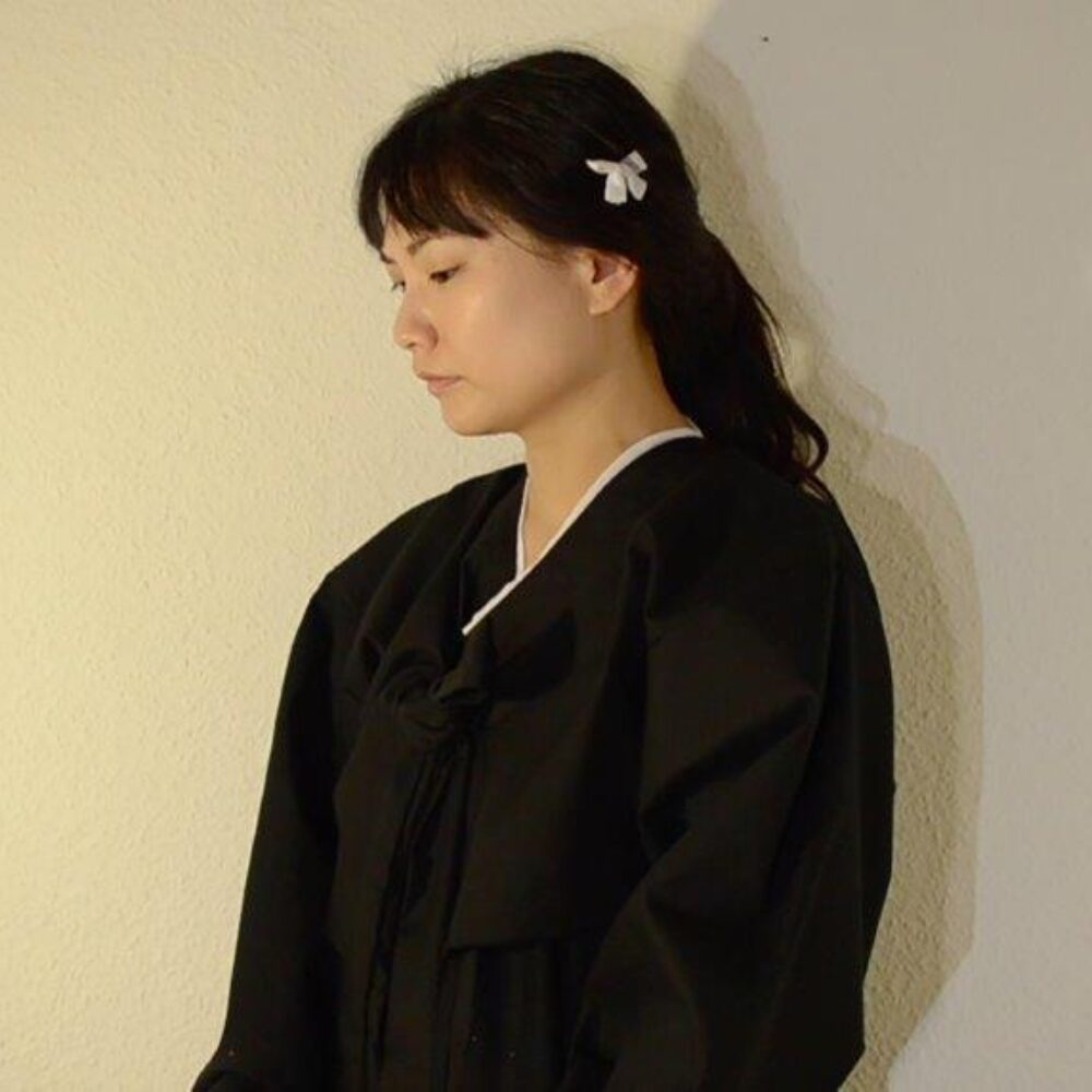 Woman with black hair tied in a braid and a black cape, standing in semi-profile in front of a white wall