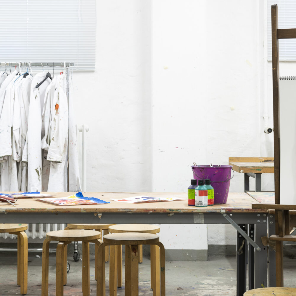 Arbeitsraum mit Staffelei mit Bild, einem Tisch mit Hockern und Bastelutensilien sowie Arbeitskittel an einer Kleiderstange im Hintergrund