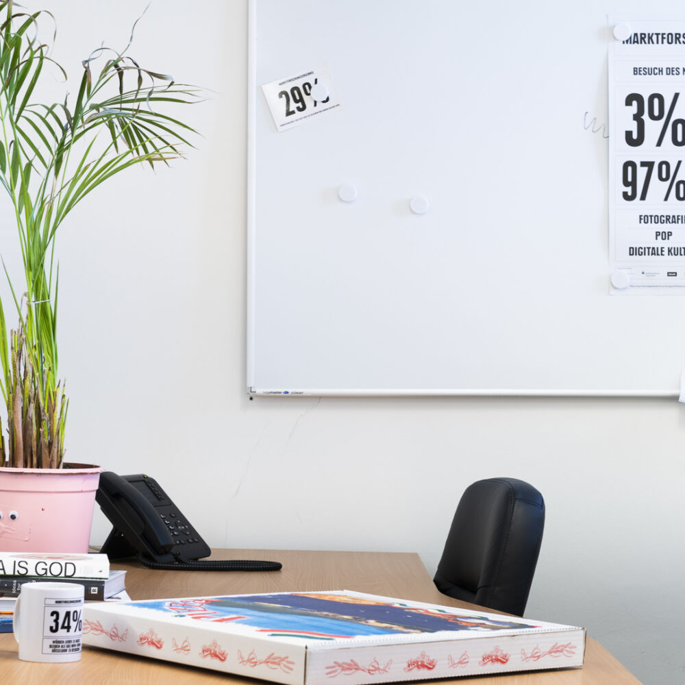 Arbeitstisch auf dem eine Pflanze, ein Telefon Bücher eine Tasse und ein Pizzakarton stehen vor einem Whiteboard an dem ein beschriftetes Poster hängt