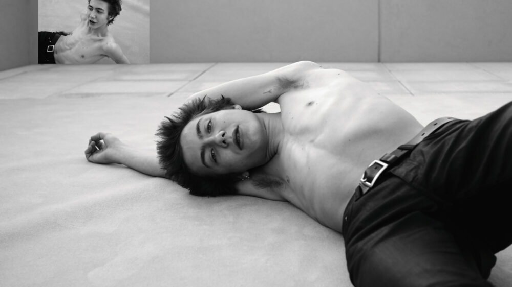 A young man with black hair is lying on the floor topless with his arms above his head