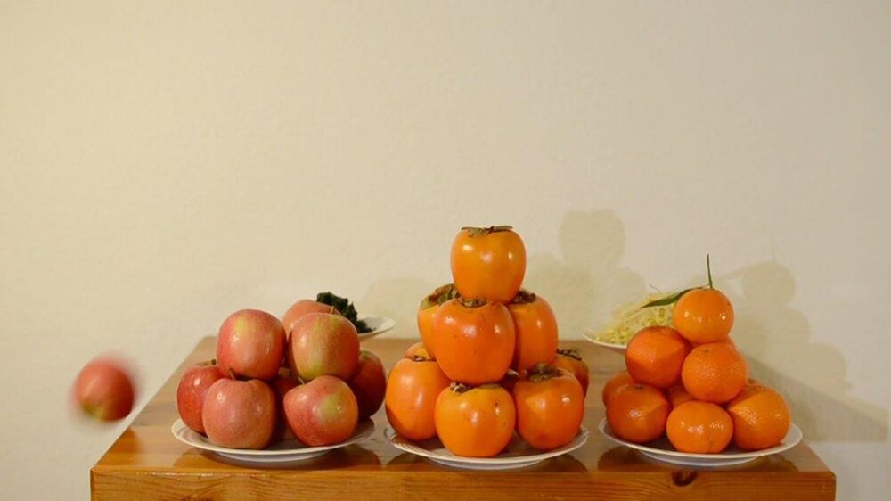 Drei Teller auf einem Tisch vor einer weißen Wand mit jeweils einem Haufen aus Kakis, Orangen und Äpfeln