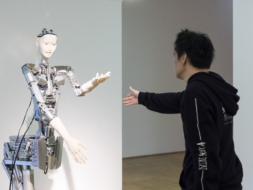 Ein schwarz gekleideter Mann reicht einem Roboter die Hand in der Ausstellung Körperwende