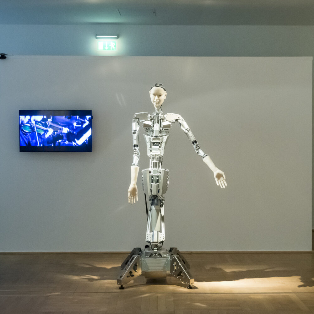 Ein Roboter steht in der Ausstellung Körperwende vor einer weißen Wand, an der ein Bildschirm hängt