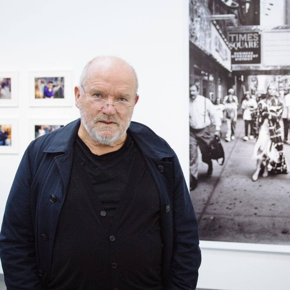 Der Fotograf Peter Lindbergh steht schwarz gekleidet in der Ausstellung Lindbergh Winogrand vor einer großen schwarz weiß Aufnahme und kleineren Fotos