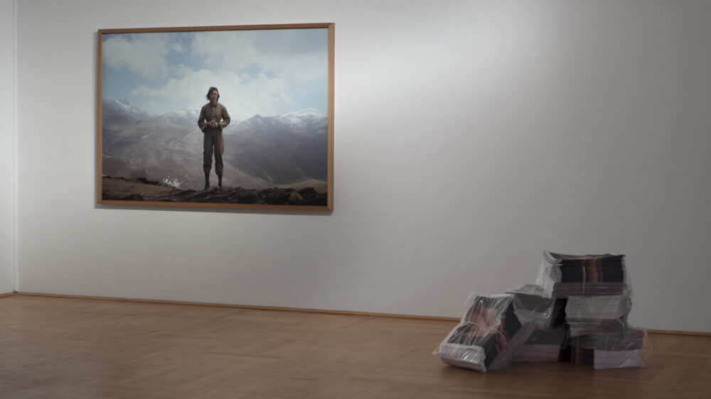 A large photo of a person hangs on a white wall and in front of it is a stack of newspapers