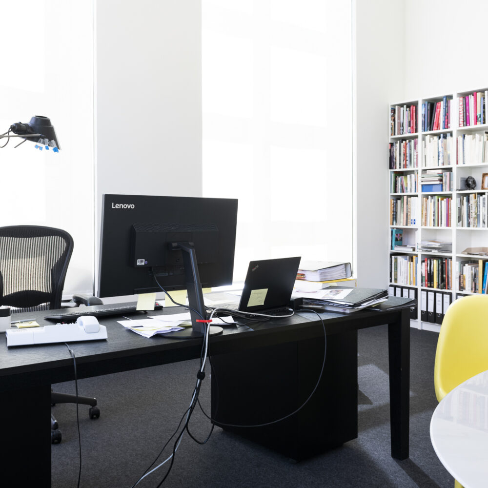 Arbeitszimmer mit schwarzem Schreibtisch mit Computer, Lampe und Unterlagen sowie einem Konferenztisch mit gelbem Stuhl und Bücherregal im Hintergrund