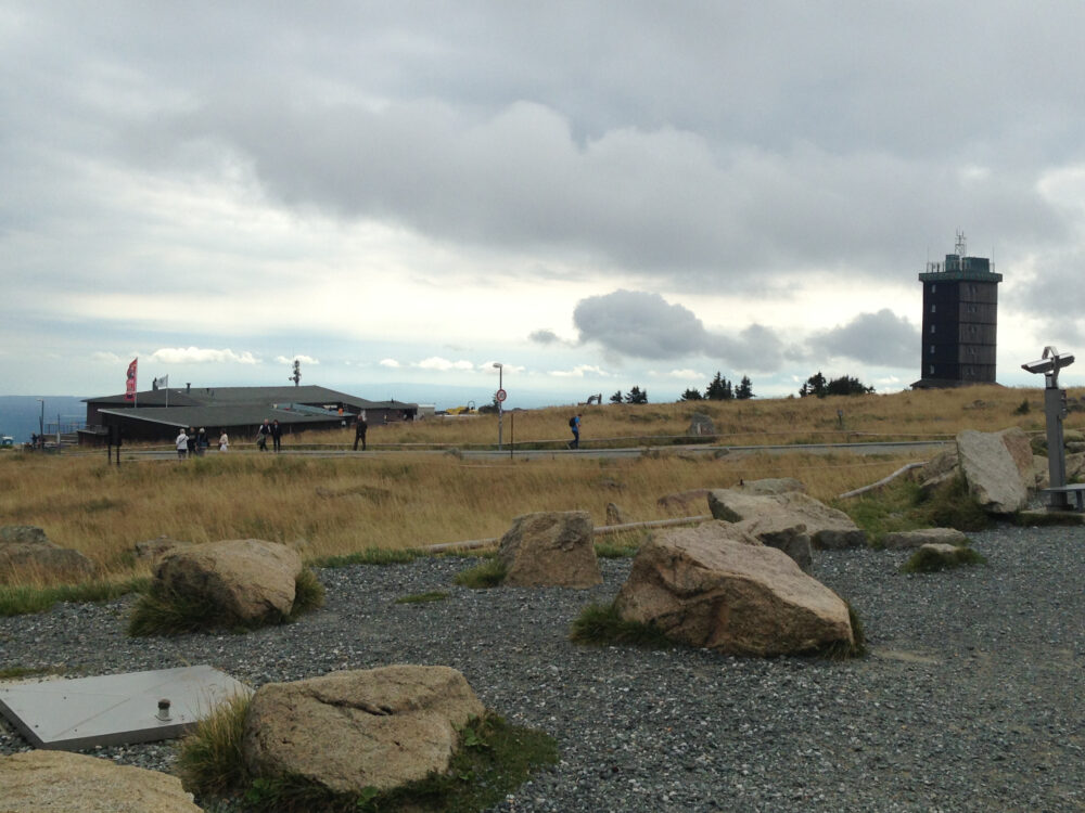 Ein Schotterweg im Vordergrund führt durch eine Wiese auf einem Berg mit einem Haus und einem Turm und einer Aussicht aufs Land im Hintergrund