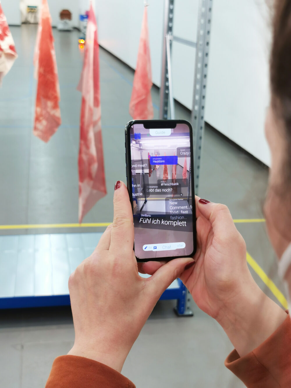 A phone is held by a woman whom we only see outlines of. There are comments on her screen, flesh looking fabric hanging in the exhibition space.