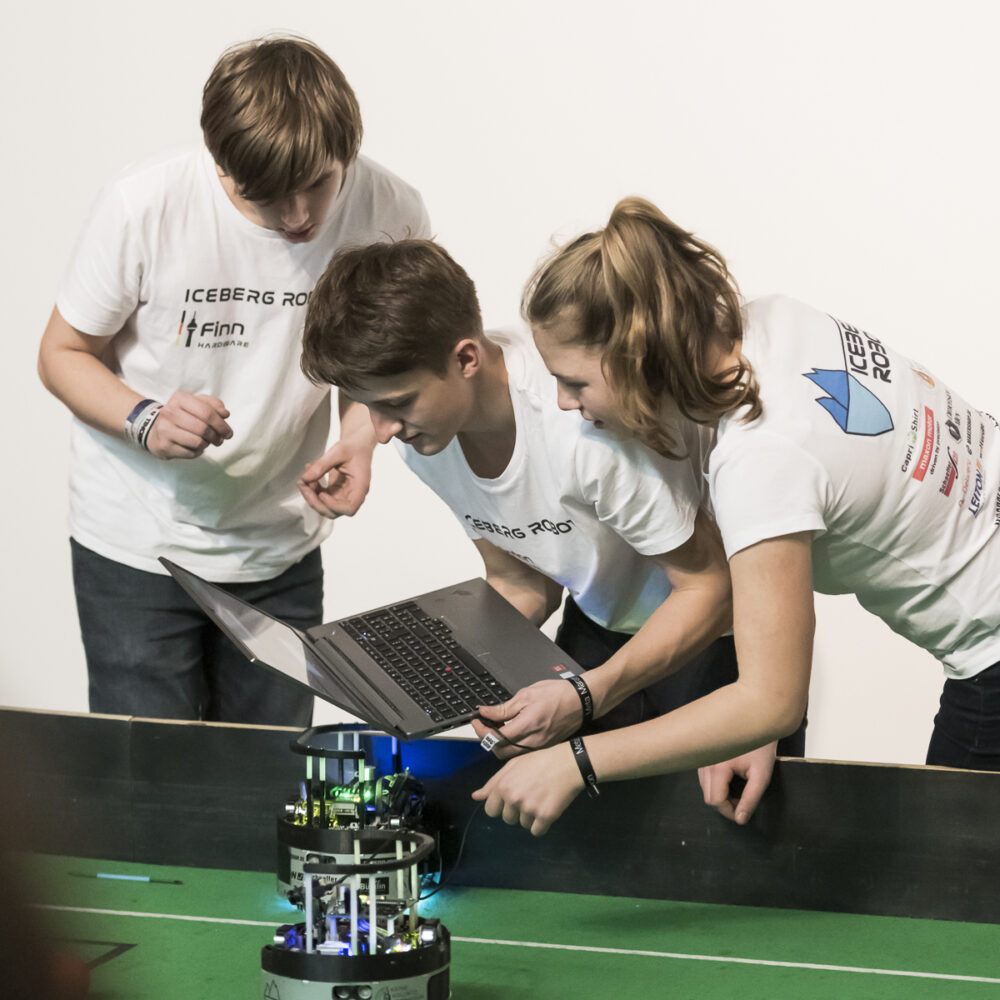 Jugendliche mit Laptop in der Hand programmieren einen Roboter, der auf einem grünen Spielfeld steht