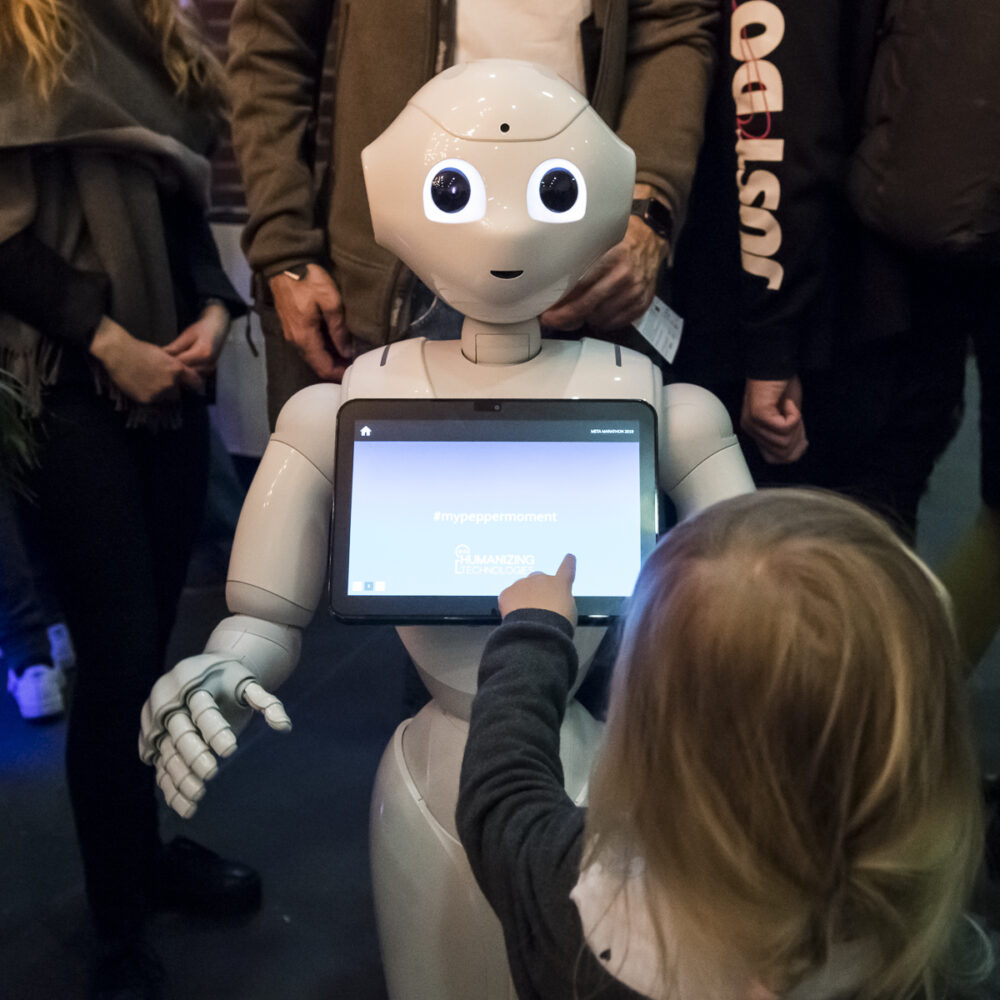 Kind bedient bei einer Veranstaltung mit vielen Leuten im Hintergrund einen weißen Roboter mit Touchscreen